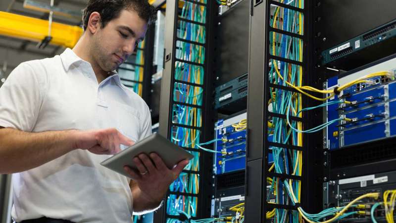 A network administrator looks at his tablet while standing in a network closet.