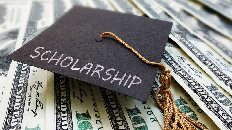 A graduation cap with the word scholarship written on it, sits on top of one-hundred dollar bills.