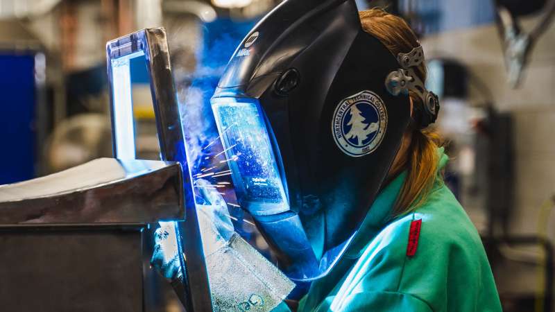 A girl is welding a piece of metal with her welding shield on as sparks fly. 