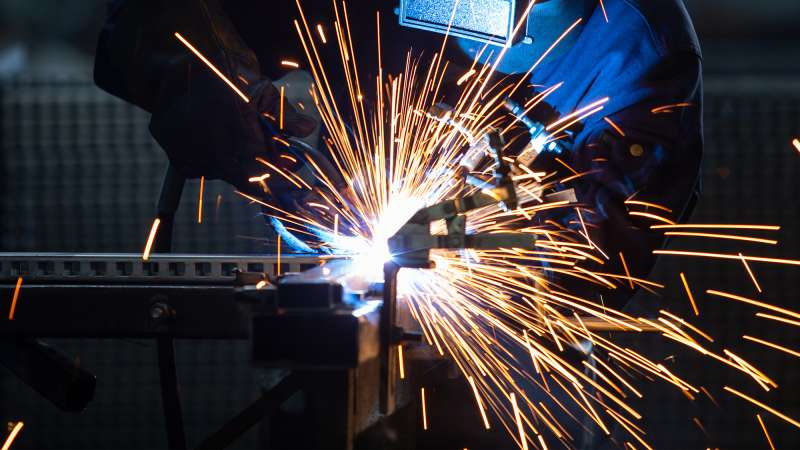 A welder wearing an industrial uniform and welding mask is welding steel inside of a factory.