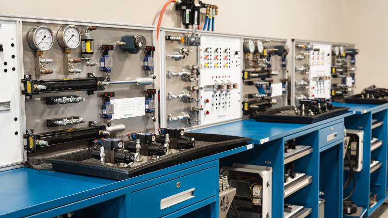 A fluid power training station featuring hydraulic and pneumatic components, including gauges, valves, actuators, and control panels. Blue workbenches support hands-on learning, with neatly arranged equipment for industrial fluid power technology training.