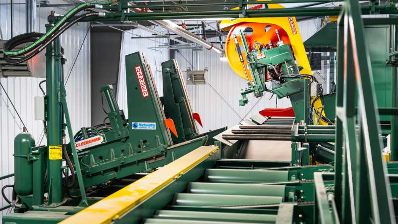 A green industrial head rig with Cleereman Industries and Northcentral Technical College branding, used in sawmill operations, is housed in a large facility. It features yellow safety components and labeled hazard warnings.