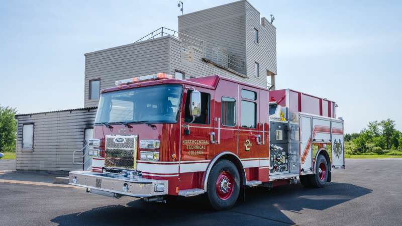 A red fire engine labeled Northcentral Technical College is parked on a blacktop surface near a multi-story training facility under a clear blue sky.
