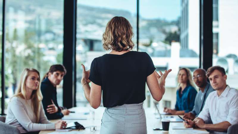 A woman stands with her back to the camera, speaking to a group of colleagues seated around a conference table in a modern office with large windows.