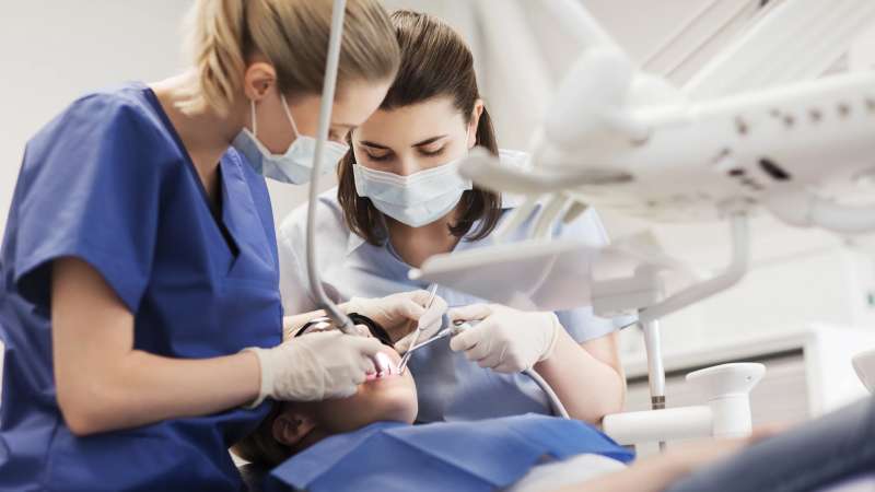 Two dental professionals wearing scrubs, gloves, and face masks performing a dental procedure on a patient lying in a dentist’s chair, with dental instruments and overhead equipment visible.