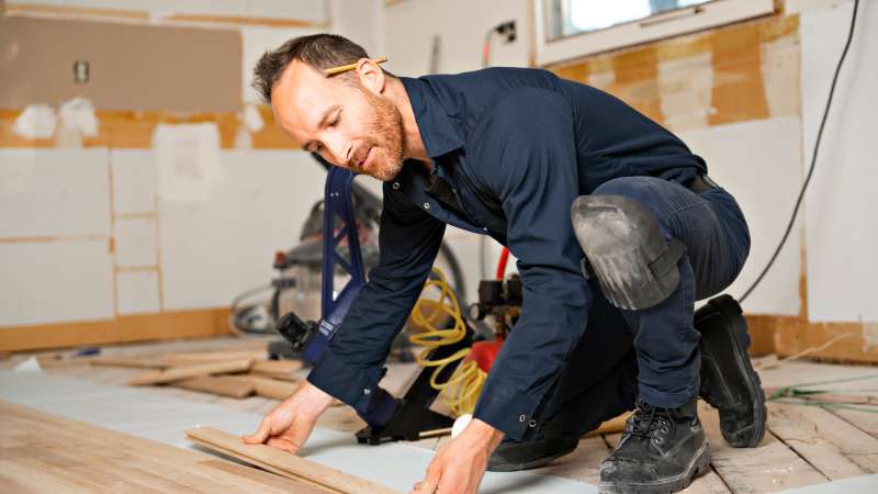 A flooring technician in work boots and kneepads installs wooden floorboards in a partially renovated room, carefully aligning each plank with tools visible in the background.