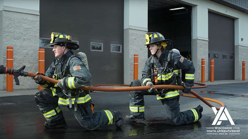 Two firefighters in gear kneel and pull a fire hose outside a fire station.