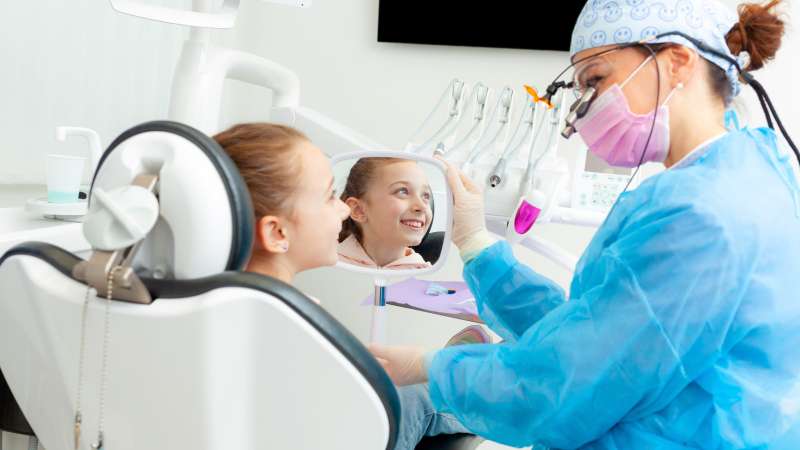 A young girl smiles at a female dentist in blue scrubs, a mask, and magnifying glasses, during a check-up in a modern dental office.