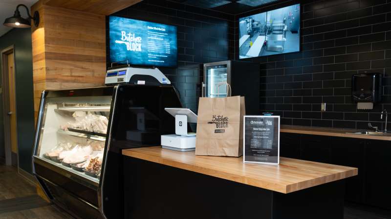 Butcher Block storefront, including shop counter with a glass display case of meats, scale, and tablet. A branded paper bag sits under two wall-mounted screens.