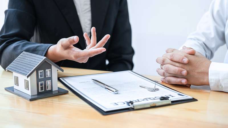 A real estate agent in a dark suit explains details to a client, with a miniature house, papers, pen, and key on the desk.
