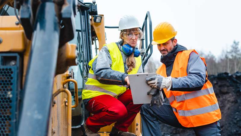 Two construction workers, a woman and a man, in hard hats and safety vests, review a clipboard together near heavy machinery.