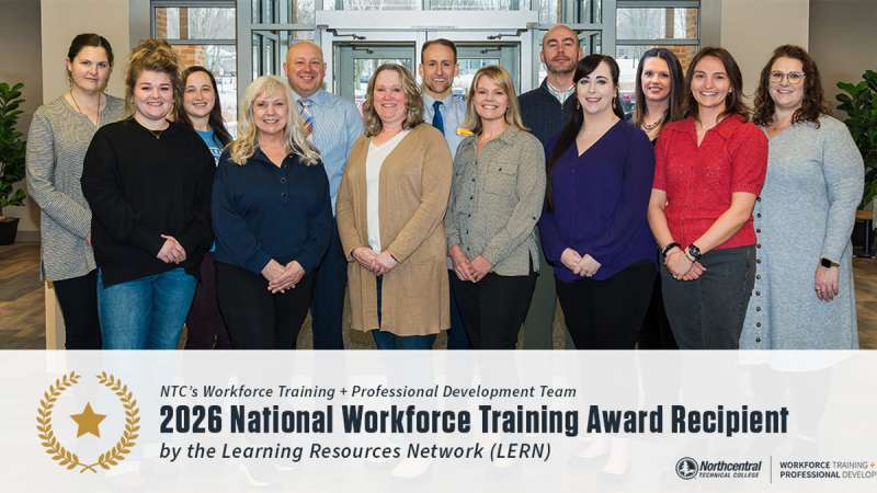 Fifteen smiling adults in business-casual attire stand in two rows in a conference room, posing as an award-recipient group.