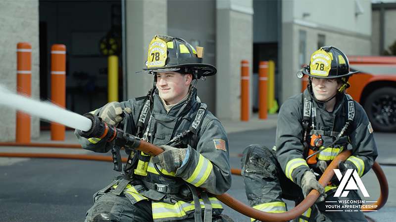Oliver Janssen and Andrew Lingl in turnout gear shoulder a hose spraying water outside a station.