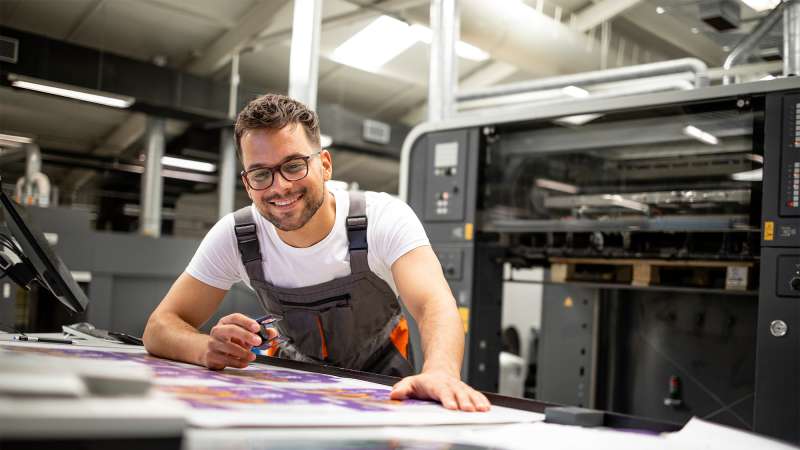 Smiling man with glasses and apron leans over a table of colorful printed sheets beside an industrial printing press.