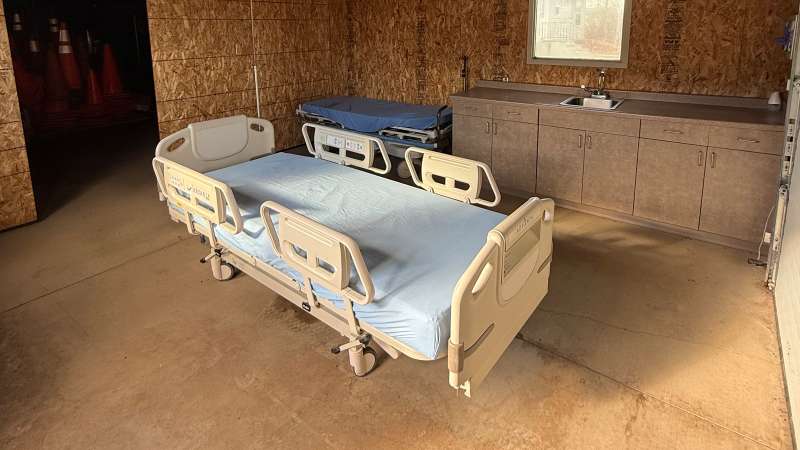 Two metal adjustable hospital beds with light blue sheets side-by-side in a small unfinished wooden room with cabinets, concrete floor, and single window.