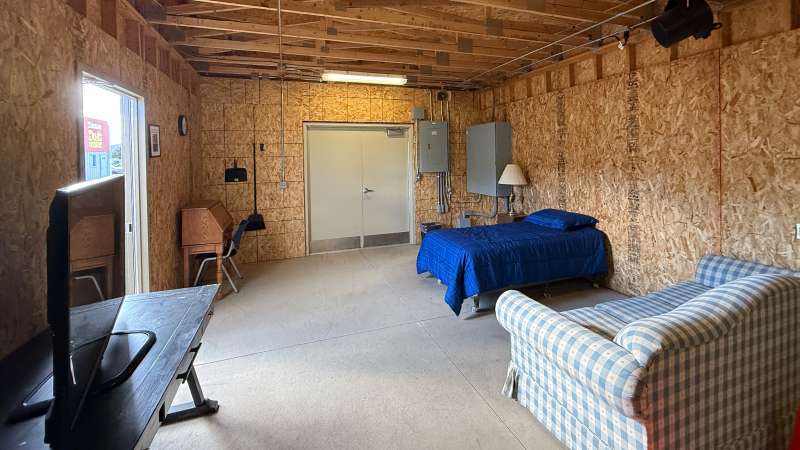 Unfinished wood-paneled room serving as a living space with bed, couch, desk with monitor, small kitchenette and window.
