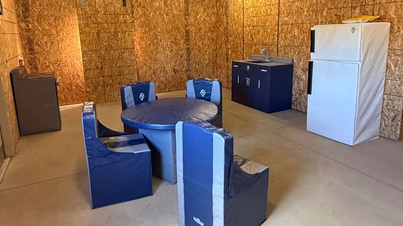 Unfinished room with OSB plywood walls and concrete floor, scattered modular blue foam seating and cabinets, plus a foam refrigerator.