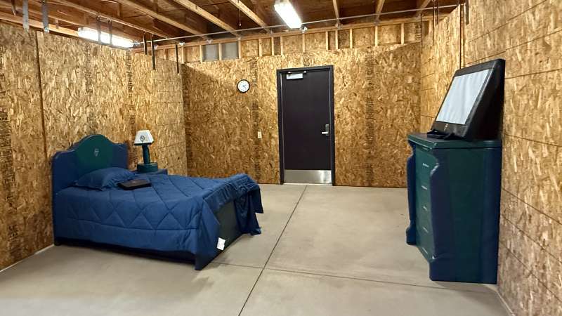 Unfinished room with OSB walls, concrete floor, exposed ceiling, furnished with a foam bed, nightstand, dresser, and TV.