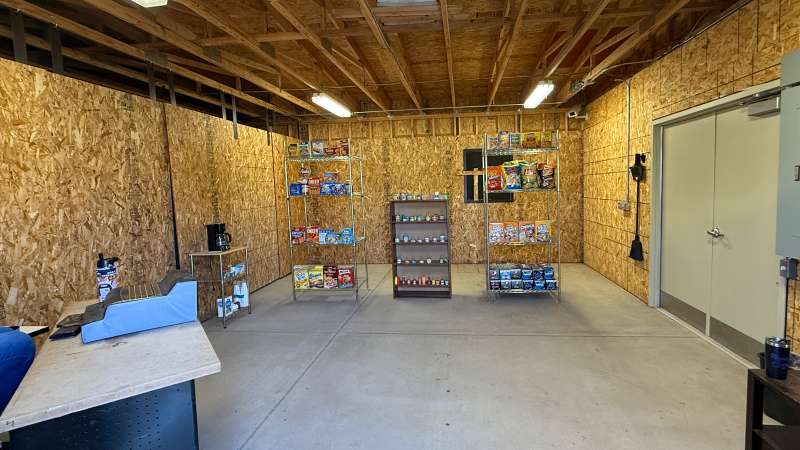Unfinished room with plywood walls and exposed ceiling beams, serving as a mock convenience store with groceries and a table.