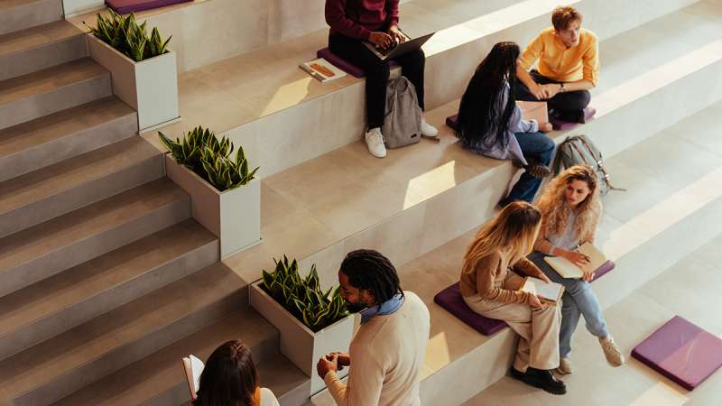 Students seated on wide tiered concrete steps in a sunlit campus atrium, reading, using laptops and chatting.
