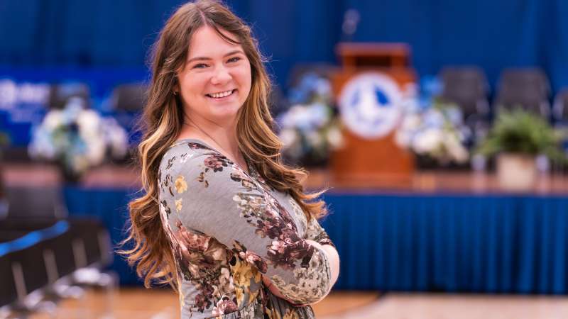 A young woman (Ashley Aull) with long wavy hair smiles while standing with her arms crossed in front of a graduation ceremony stage decorated with flowers and a podium. She wears a floral dress and poses in a gymnasium setting prepared for commencement activities.