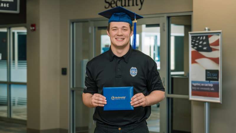Kyle smiling while wearing his graduation cap and holding up his diploma in front of the Safety and Security Office.