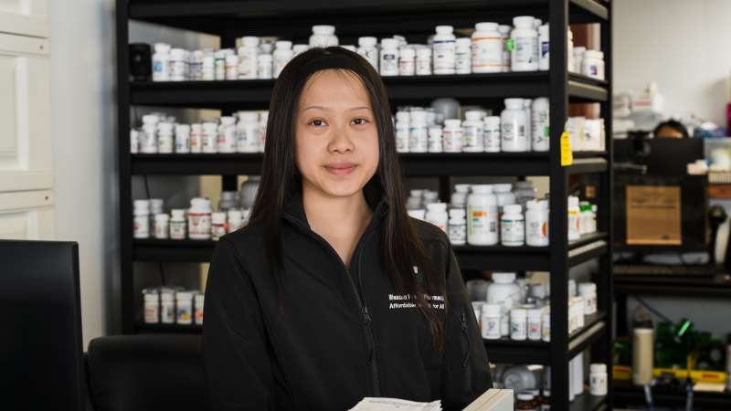 Nancy Xiong, a pharmacy technician youth apprentice, stands behind a pharmacy counter, looking toward the camera, with shelves of prescription medication bottles neatly organized behind her. She wears a black pharmacy-branded jacket and is positioned in a working pharmacy environment that includes medication storage, dispensing supplies, and computer workstations.