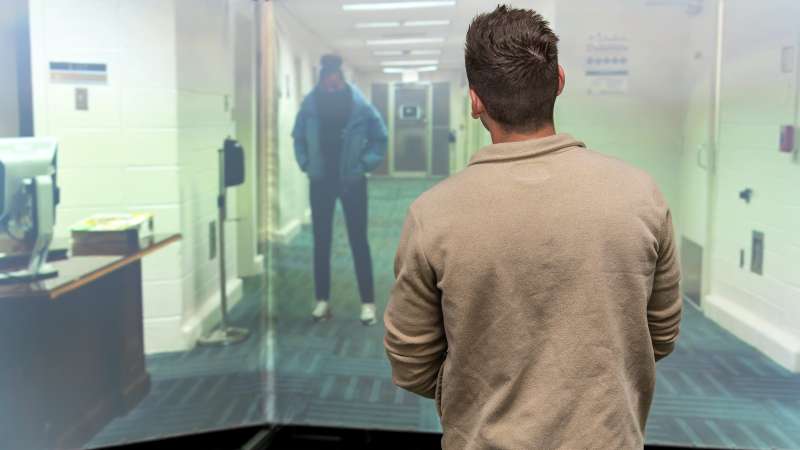 Person seen from behind interacts with a life-size projection of a person standing in a school hallway.