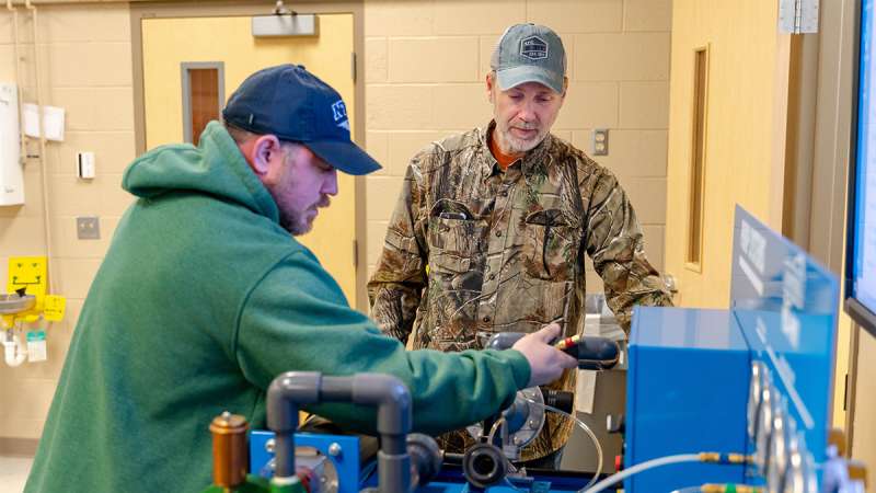 Two men focused on metal machinery in a workshop: one in a green hoodie adjusting equipment, another in camouflage jacket.