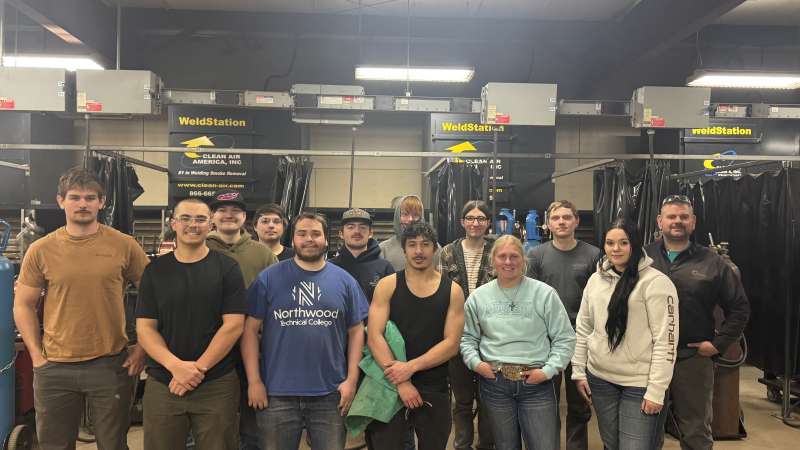A group of students and an instructor pose together. The students stand in front of a row of welding booths equipped with ventilation systems and welding curtains, representing a variety of technical training backgrounds. Industrial equipment, welding stations, and safety infrastructure fill the workspace behind them.
