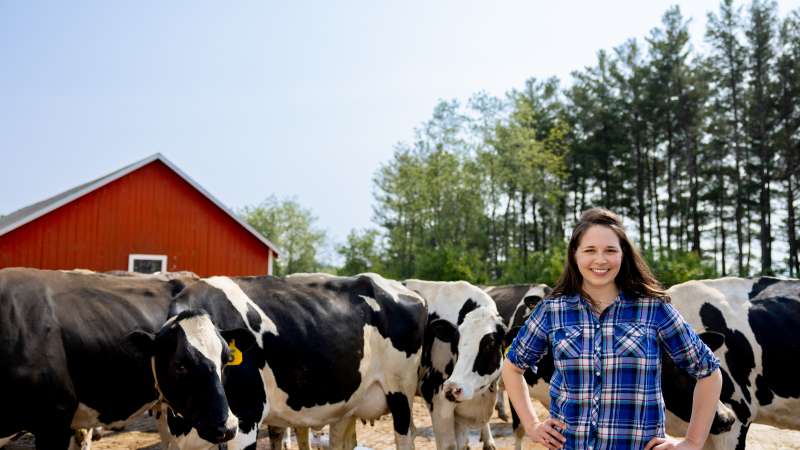 A photo shows a person standing with hands on their hips beside a group of black-and-white cows. A red barn and a line of trees are visible in the background.