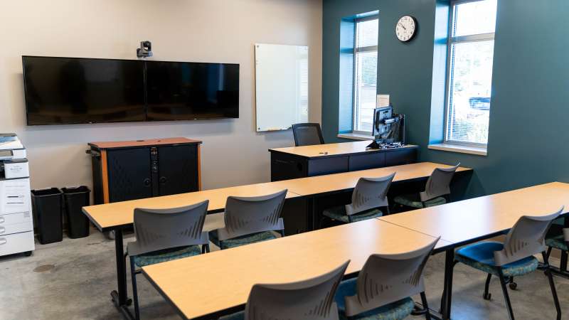 Training room with three rows of tables and gray chairs facing a large flat-screen monitor, whiteboard, clock, and windows on a teal wall.