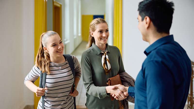 Smiling female teacher in green blazer shakes hands with a man in a school hallway with a smiling girl with a backpack.