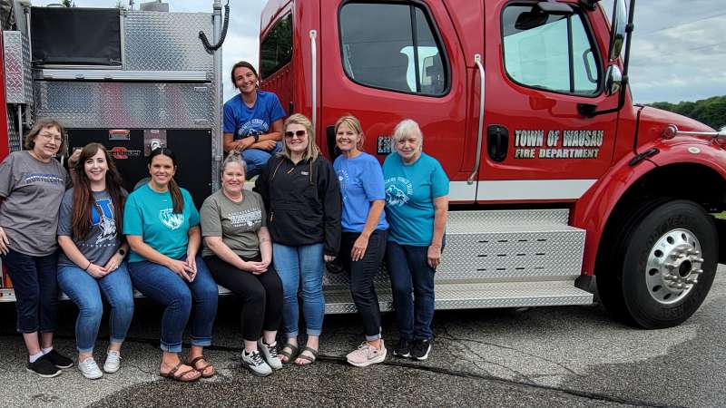Group photo of NTC employees smiling and posing beside a red Town of Wausau Fire Department truck during a community service activity. Several employees wear NTC Timberwolves shirts in blue, teal, gray and purple.
