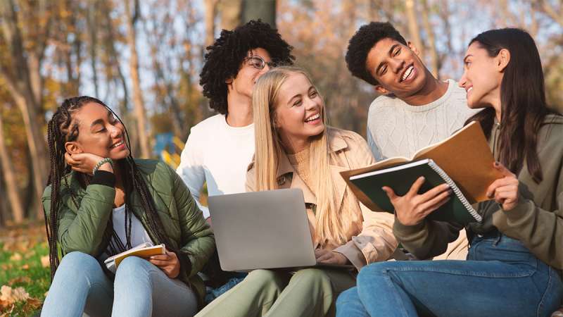 Five diverse college students sit on grass in an autumn park, laughing and studying together with notebooks and a laptop.