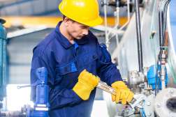 A student wearing a blue jumpsuit and a yellow hardhat is tightening a bolt with a wrench