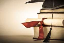 A graduation hat sits on top of three large books and a rolled up paper wrapped in a red bow