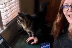 Laura working from home with her cat sitting on her desk next to her.