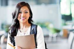 A student stands in a classroom with a pencil in hand and a notepad in her arms.