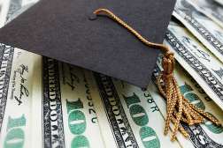 A black graduation hat with gold tassels sits on top of several one-hundred dollar bills.
