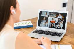 A woman using earbuds and a laptop at a desk, is attending an online meeting with several other people.