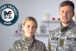 Two military soldiers stand side by side in uniform with the American flag behind them.