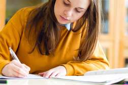 A young college student in sits at a table with open books writing notes down. 
