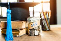A graduation hat is placed next to a jar full of pencils, a couple of textbooks, and a jar full of coins.