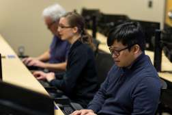Three students in a classroom are typing on keyboards at desktop workstations.