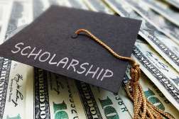 A graduation cap with the word scholarship written on it, sits on top of one-hundred dollar bills.