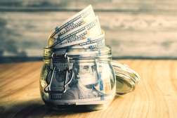 An open jar on top of a wooden table filled with one-hundred dollar bills.
