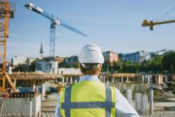 A civil engineer wearing safety gear looks over a construction site full of workers and construction equipment.