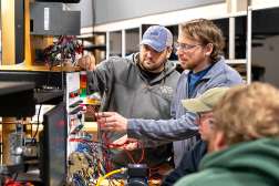 Three apprentices are being instructed as they work on electrical equipment.