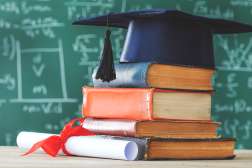 A stack of books underneath a graduate hat in front of green chalkboard with math written on it.